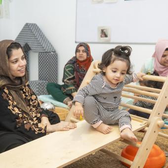 A mother looks at her daughter, playing on a Pikler learning structure.