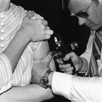 L’instrument utilitzat pel metge a la foto rebia el nom de «pistola de la pau», pel seu paper important en campanyes globals d’eradicació de la verola. Aquests dispositius utilitzaven aire comprimit a alta pressió per disparar un raig de vacuna tan fi que travessava la pell sense necessitat d’utilitzar agulla.